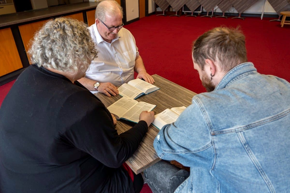 Three adults seated around a table in a parish hall, each with an open Bible, reading together.
